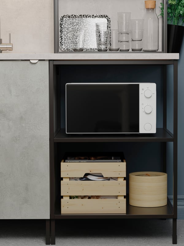 A microwave and two wooden boxes are on open shelves under a kitchen worktop. Beside is a grey cabinet with door.