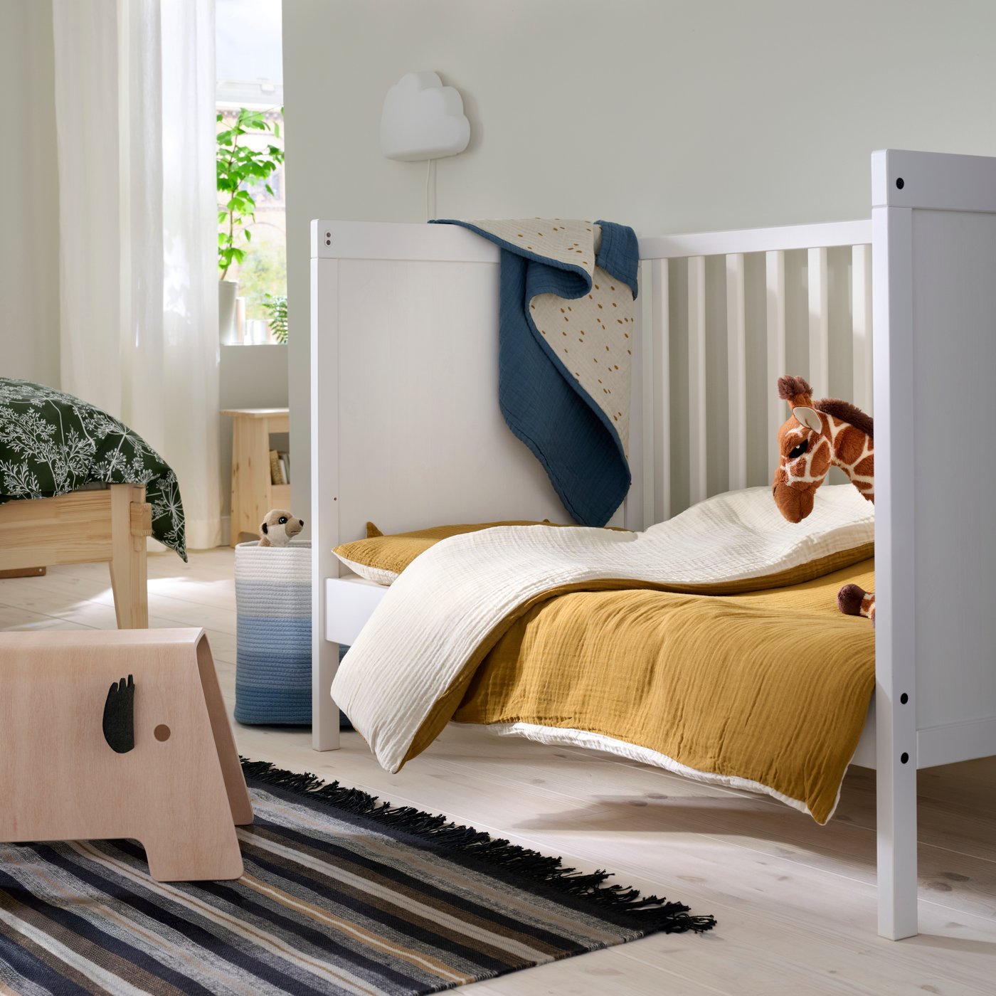 Una cuna SUNDVIK en un dormitorio familiar con un edredón marrón y un peluche de jirafa.