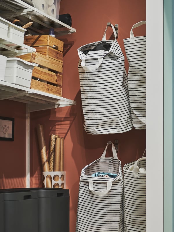 Quatre sacs à linge KLUNKA en noir et blanc suspendus à des crochets muraux, à côté de tablettes sur lesquelles sont posées des boîtes.