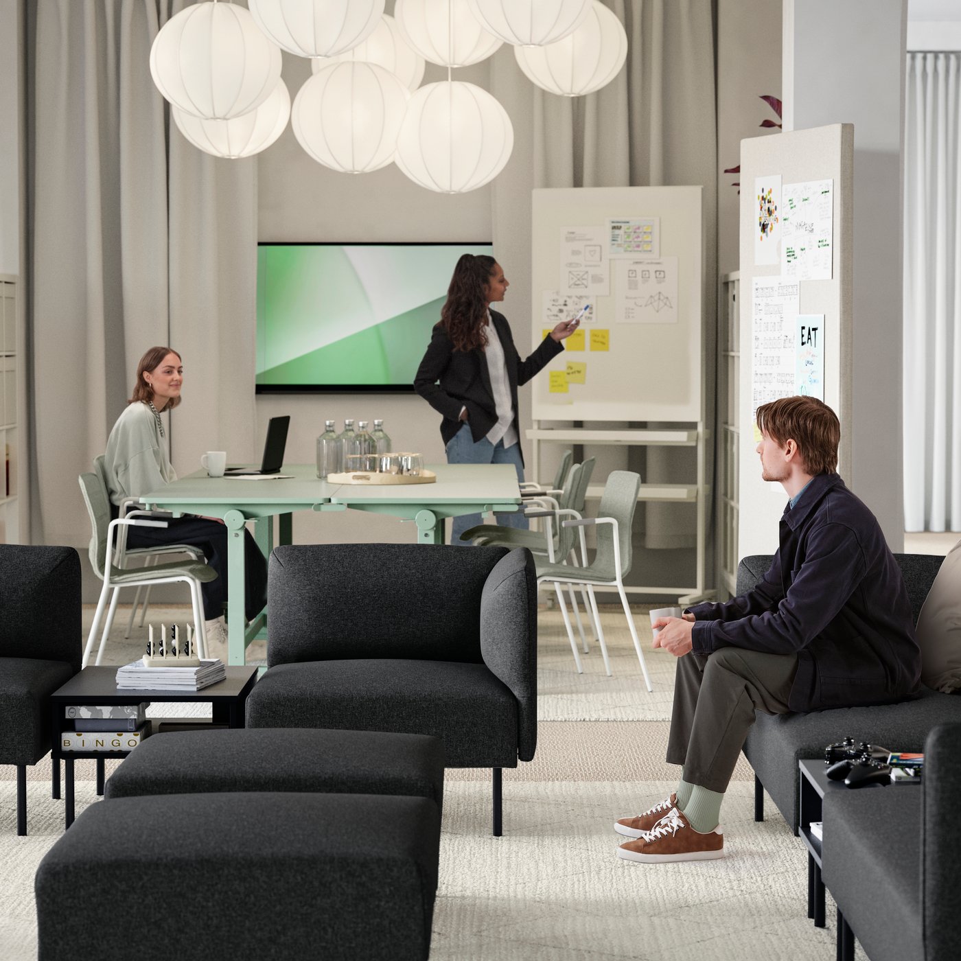 A young man sitting in a lounge area of several dark grey LILLEHEM sofa modules, TUNSTA tables and a meeting area behind it.