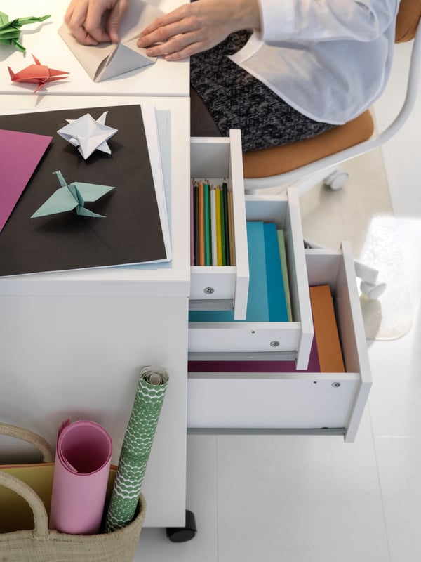 Una cajonera MICKE blanca con tres cajones abiertos mostrando papeles de diferentes colores en su interior. Al costado una jove haciendo origami.