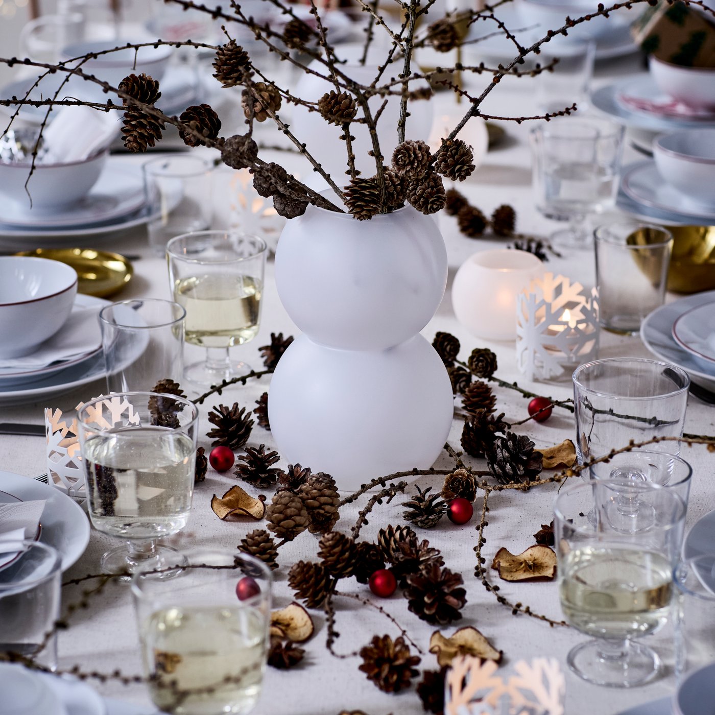 A white-themed dinner table is decorated for Christmas with a VINTERFINT vase with branches in it as a centrepiece.