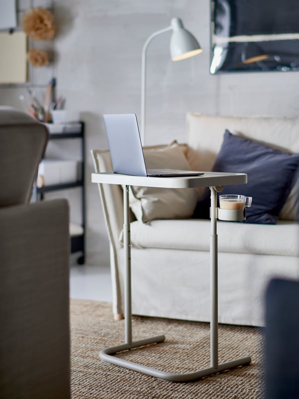 A laptop on a beige BJÖRKÅSEN laptop stand, a jute rug, an armchair with dark grey cushions and a white floor/reading lamp.