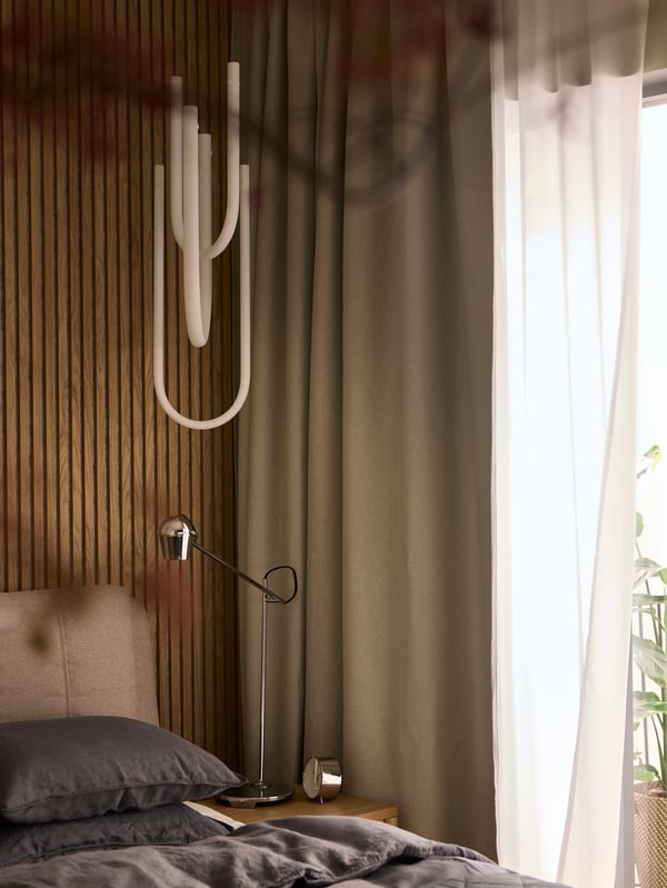Es ist die Ecke eines Schlafzimmers mit einem Fenster zu sehen, an dem abdunkelnde ANNAKAJSA Gardinen und ROSENROBINIA Gardinenstores hängen.