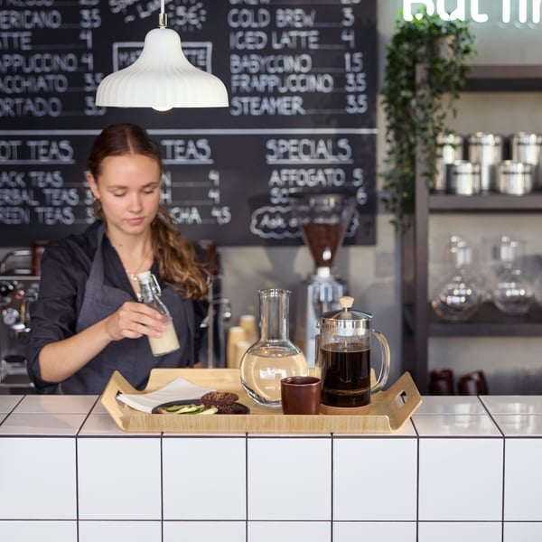Café scene with a barista preparing a bamboo SILVERHAJMAL tray to serve drinks and food on a tiled café counter.