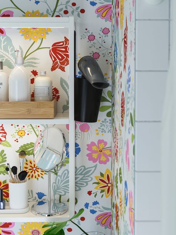 A black container with a black hairdryer hangs on the side of a white open shelf, against colourful floral wallpaper.