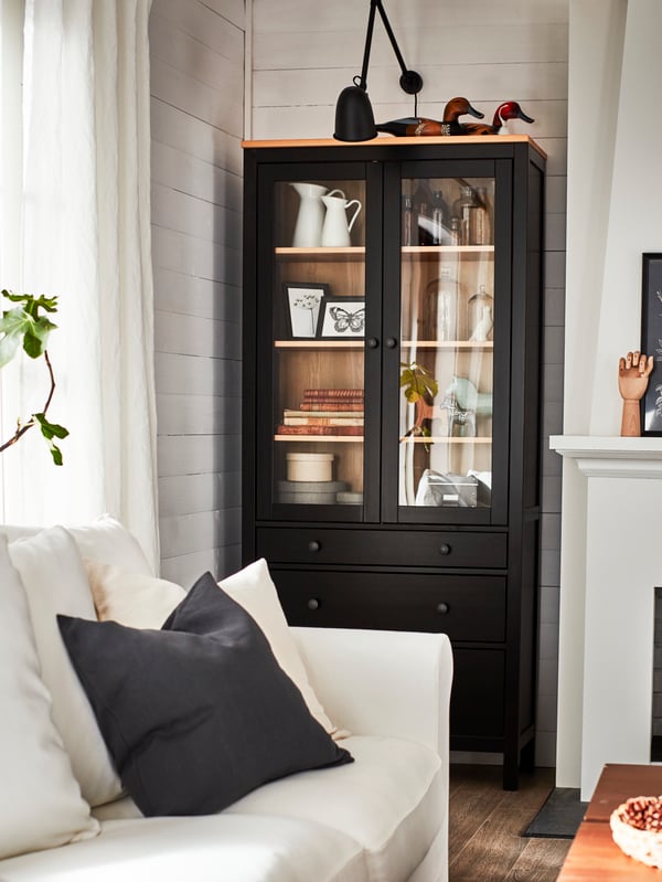 Ein schwarzbrauner Vitrinenschrank mit dekorativen Gegenständen, u. a. mit einer schwarzen Wandleuchte und einem weissen Sofa.