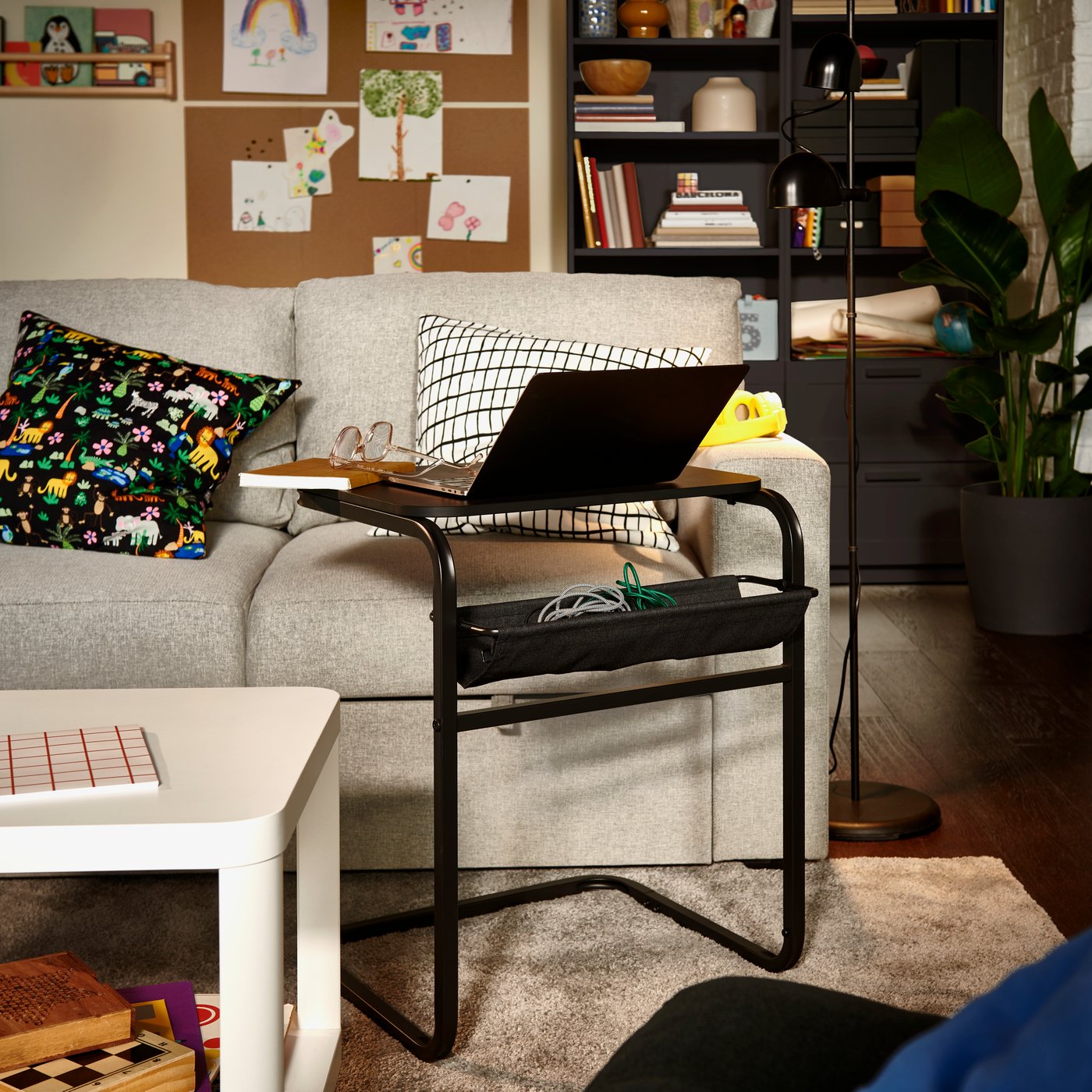 A living room with an anthracite/dark-grey OLSERÖD side table with a laptop set beside a grey sofa with patterned cushions.