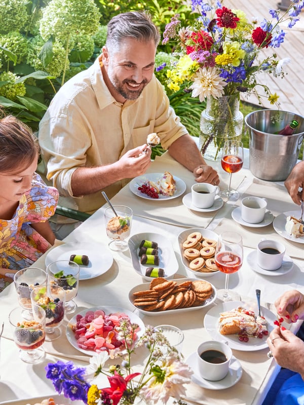 A family celebrating Swedish Midsummer outdoors, gathered around a dining table with many snacks, food and coffee served for everyone.