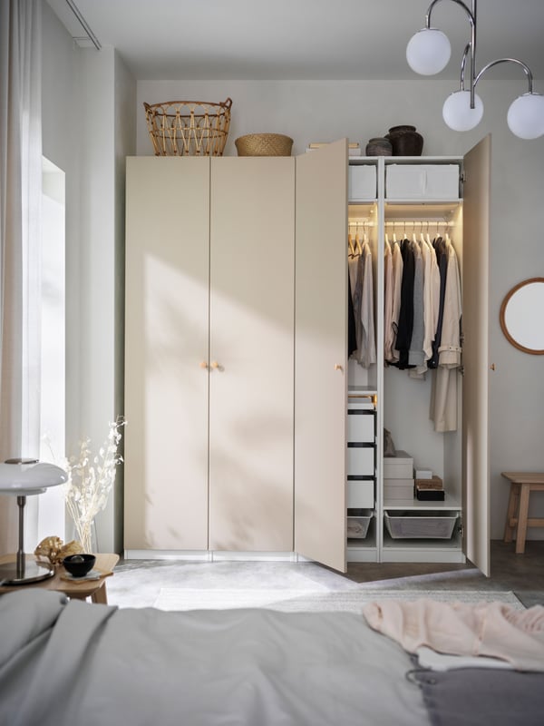 A PAX/REINSVOLL wardrobe, with clothes hanging inside it and a rattan SNIDAD basket on top of it, stands in a bedroom.