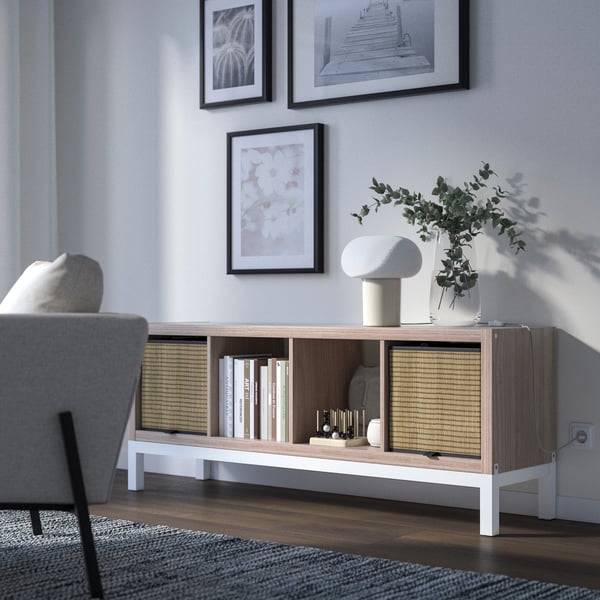 An open sideboard in which baskets, books and decorations can be seen. On the sideboard there is a table lamp and a vase, above which pictures are hung on the wall.