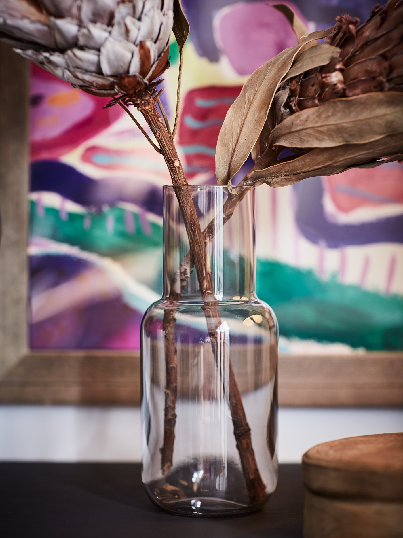 A close-up of two thick-stemmed dried flowers in a glass vase with a colourful painting in the background.