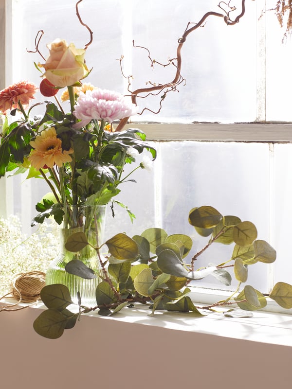 Des feuilles artificielles SMYCKA et des fleurs dans un vase sur un appui de fenêtre baigné de soleil.