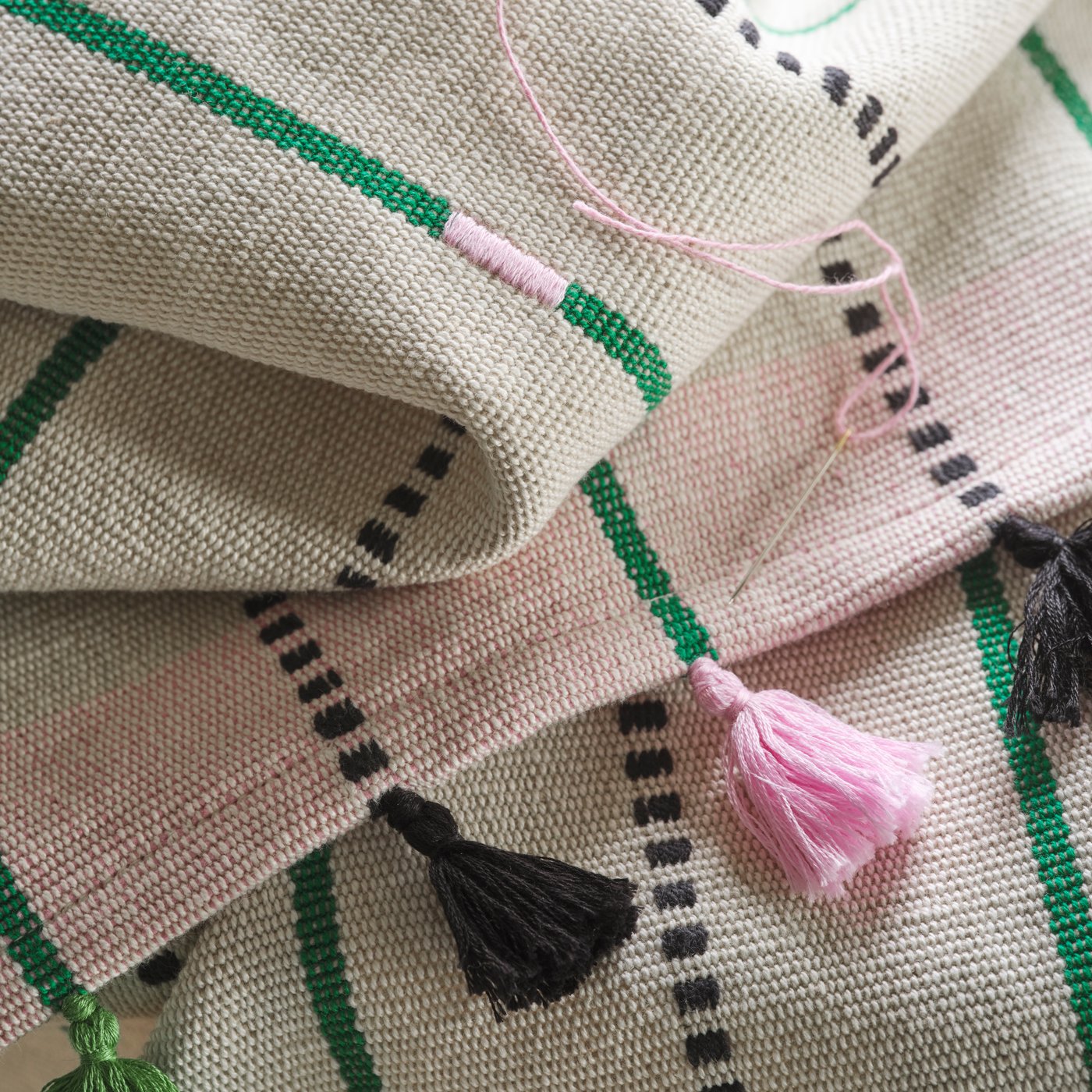 A close-up of the black and pink tassels of a handmade MÄVINN table-runner with stripes. On top is a needle with pink thread.