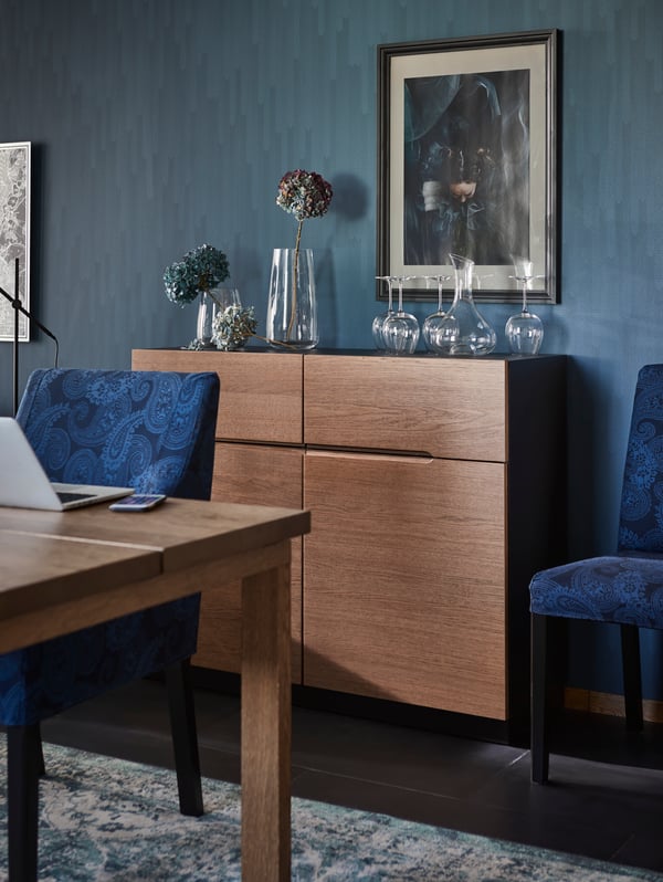 A brown KLACKENÄS sideboard against a blue wall with glass vases on top, some with flowers in, and a blue chair beside it.