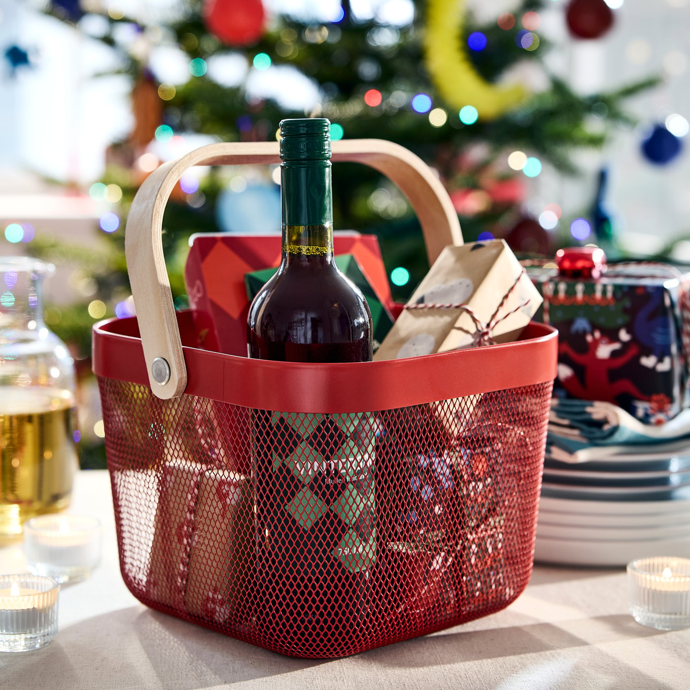 A red RISATORP basket filled with wrapped gifts and a bottle of VINTERSAGA mulled fruit drink and a gingerbread house.