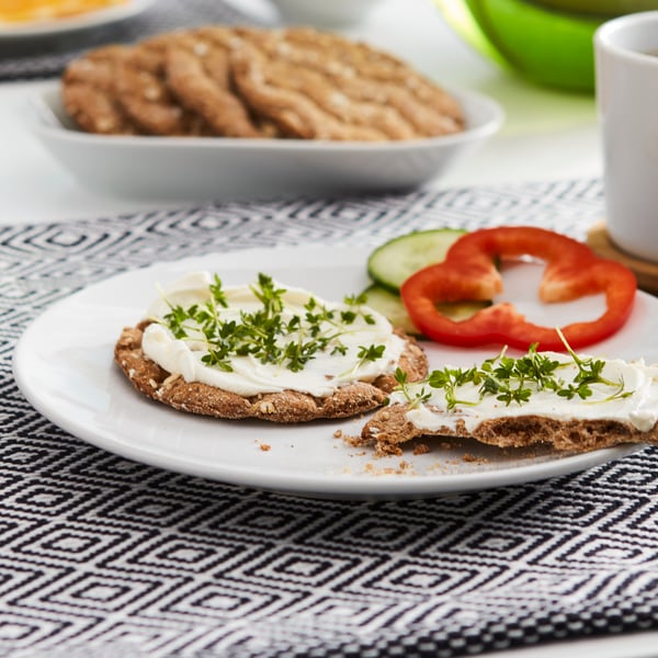 KNÄCKEBRÖD FLERKORN multigrain crispbread with creame cheese and herbs are placed on a white plate on a patterned placemat.