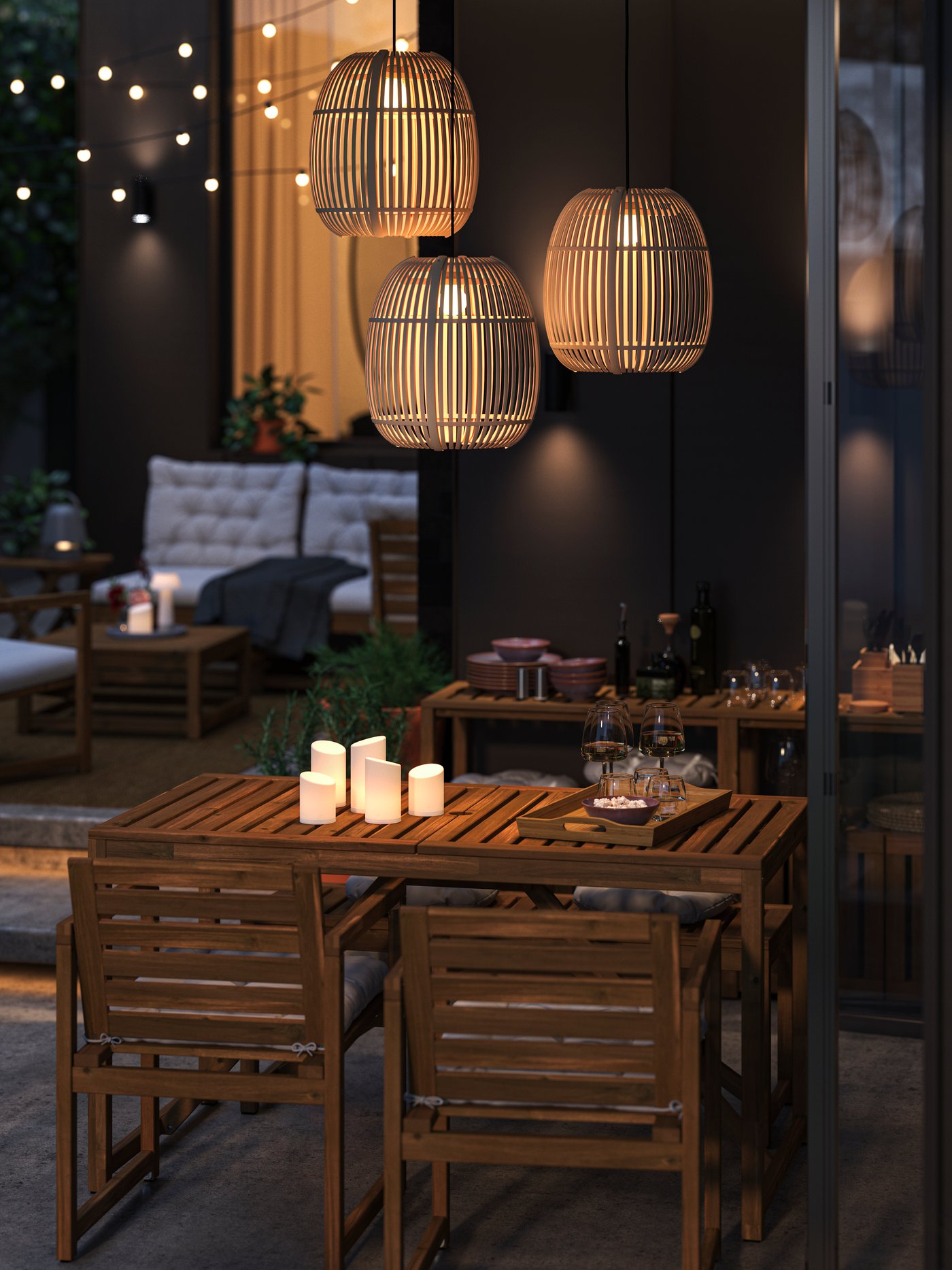 A patio dining area at dusk with three lit SOLVINDEN pendant lamps over a NÄMMARÖ outdoor table with LED candles and chairs.