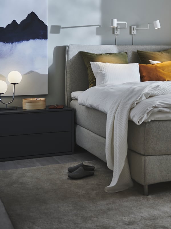 Un letto con biancheria bianca, cuscini gialli e verde scuro. Sopra, lampade da parete bianche e accanto una cassettiera - IKEA