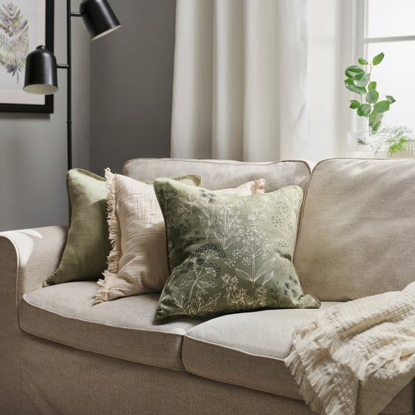 Een comfortabele IKEA-bank met groene en crèmekleurige sierkussens, een zachte gebreide plaid, zwarte wandlampen en een groene kamerplant staat in een rustig, licht interieur.