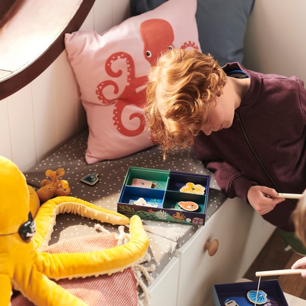 A boy plays with a BLÅVINGAD fishing game on a window seat with a cushion in a BLÅVINGAD octopus cushion cover.