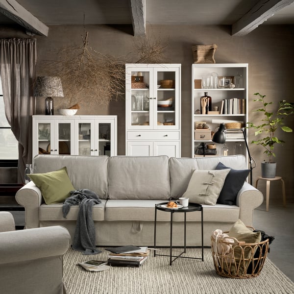 Ljust vardagsrum med beige soffa på en ljus handvävd matta, omgiven av vitrinskåp, öppna hyllor och naturinspirerade detaljer i varma toner.