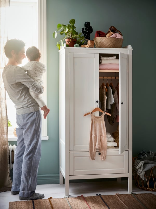 A father standing while holding a toddler with a white children's wardrobe.