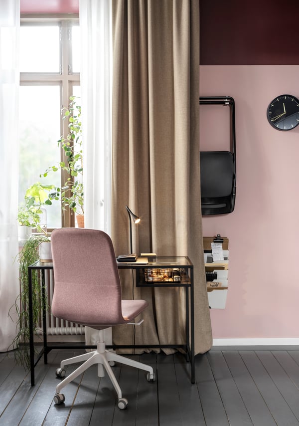A laptop table in black-brown/glass and a light brown-pink conference chair stand by a window with beige and white curtains.
