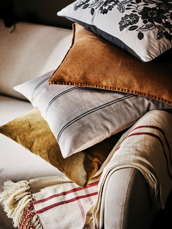 Four scatter cushions, one with a STORTIMJAN cover, on top of one another on the corner of a beige sofa with a throw.