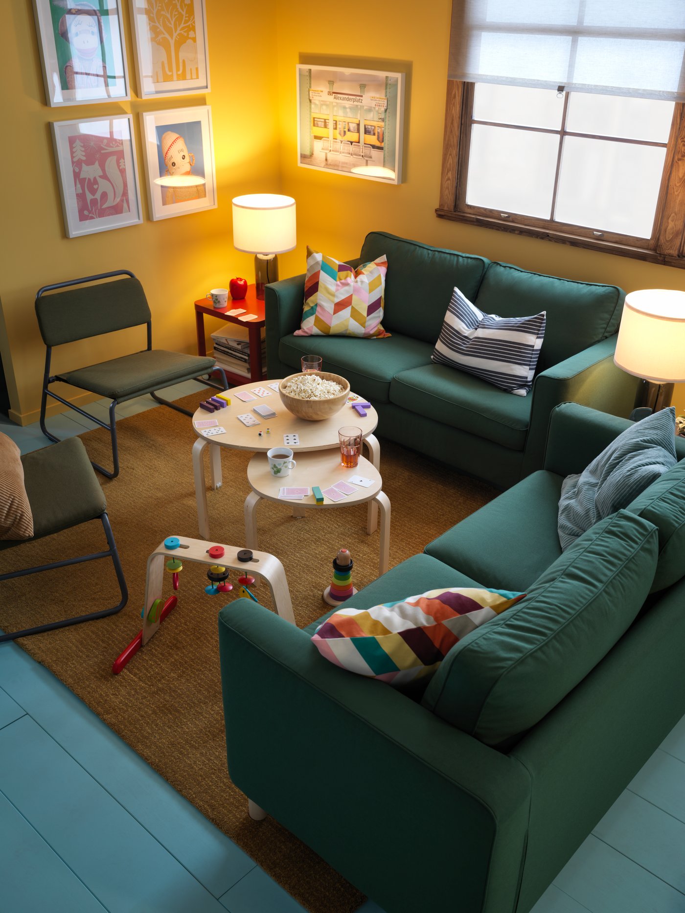 A living room corner with a pair of PÄRUP dark-green two-seat sofas framing a LEKA baby gym and SVALSTA birch nesting tables.
