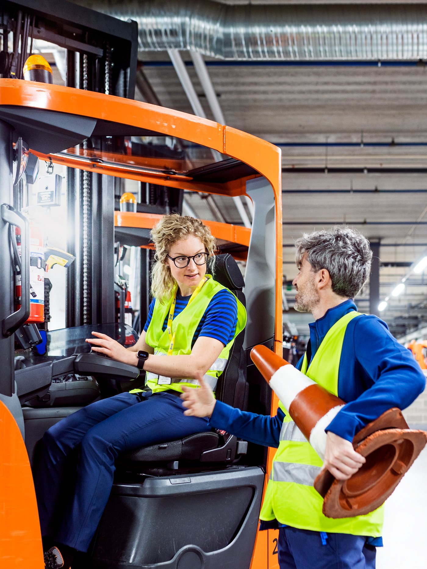 Twee IKEA medewerkers zijn aan de praat in een magazijn, een zit op een vorklift en de andere staat ernaast en houdt een verkeerskegel vast.