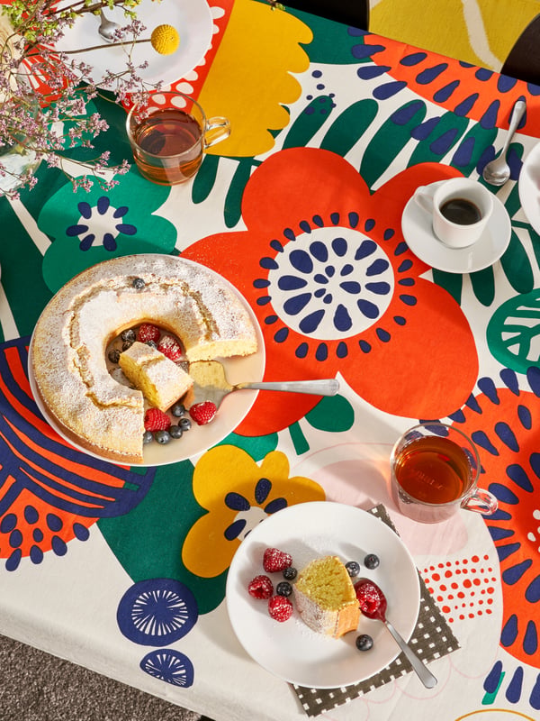 A table that has a bright flower-patterned tablecloth, plates, glasses, cups and a cake on it.