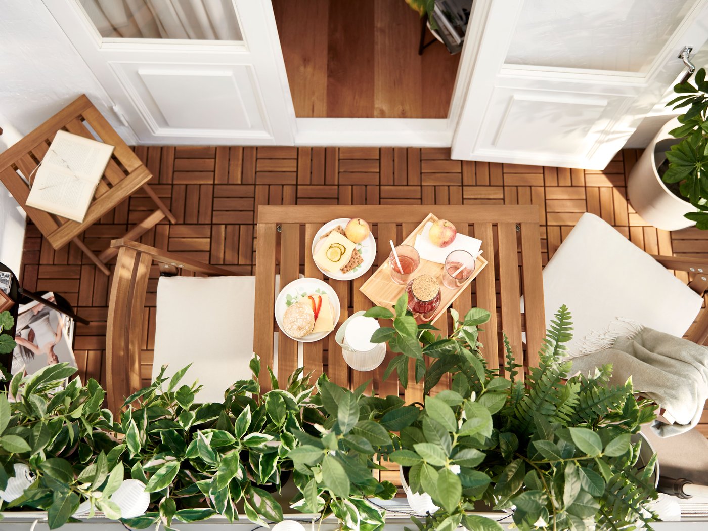 A top-down view of a balcony with wooden flooring, set for an outdoor meal. A table is laden with food and drinks, surrounded by chairs and lush green plants, creating a serene and inviting atmosphere.