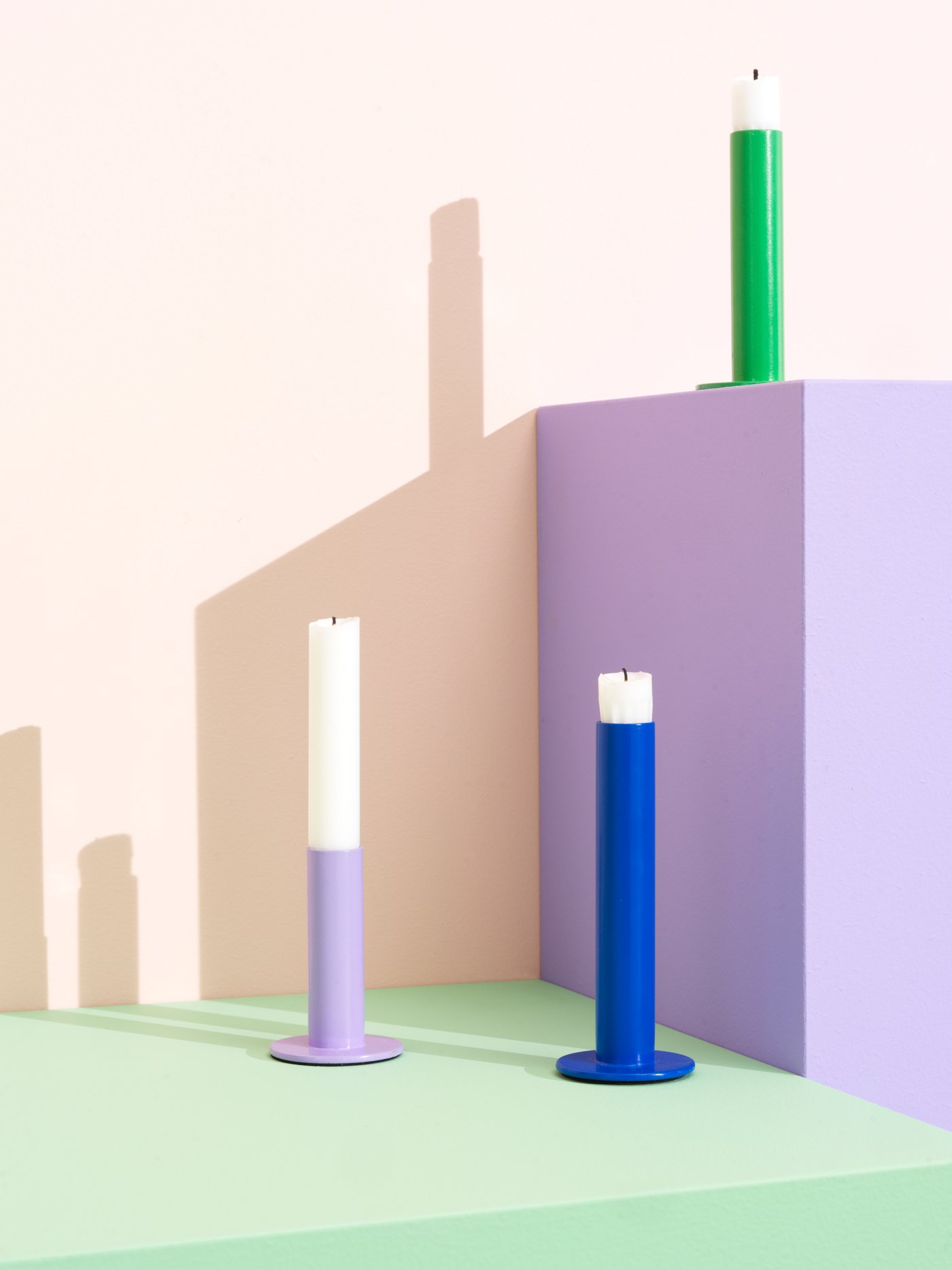 Two TUVKORNELL candle holders in lilac and blue are on a light green surface. A blue candle holder is on a lilac podium.