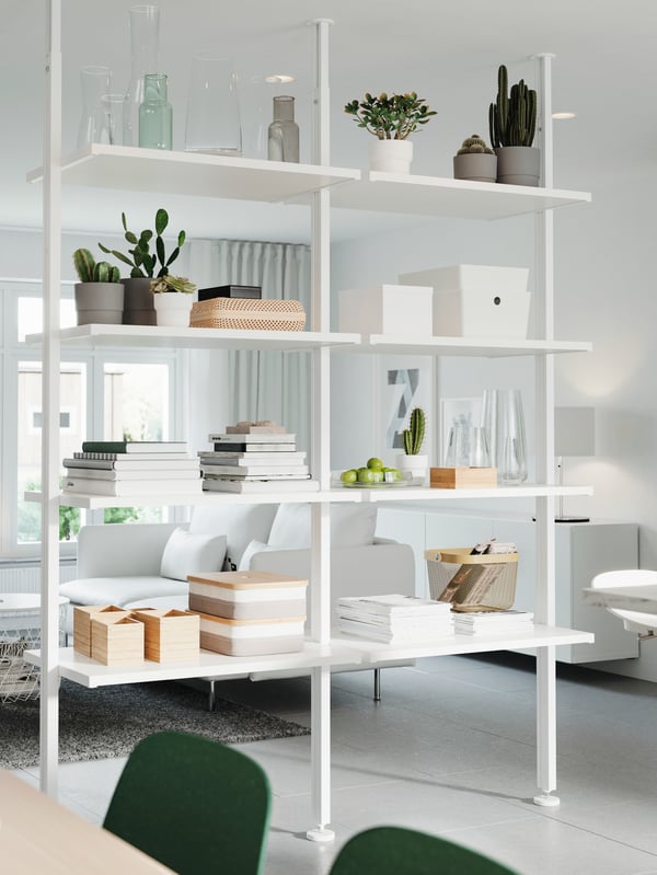 A white storage system that’s used as a room divider. Decorative items, plants, books and storage boxes stand on the shelves.