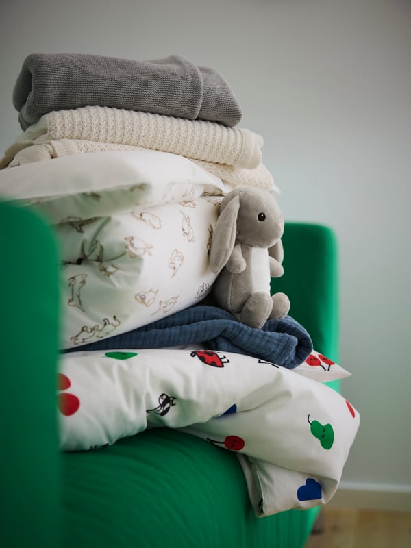 Pile de literie sur un lit bébé : couettes, oreillers, couvertures et jetés. À côté, une peluche DVÄRGHARE.
