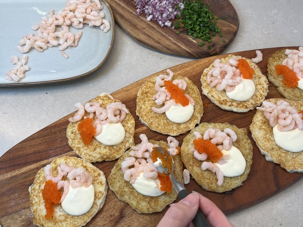 Nästan klara potatisrösti ligger på en skärbräda och toppas med räkor, kaviar och creme fraiche.