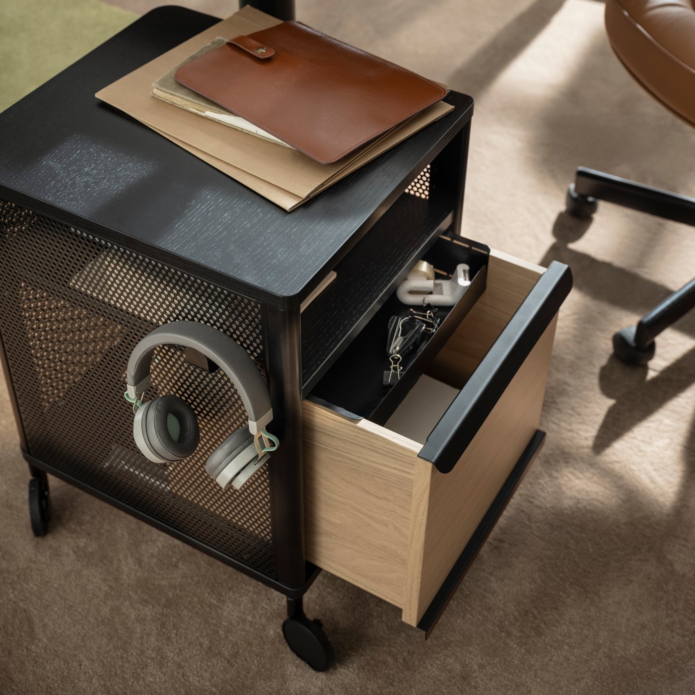 Headphones and other office accessories in and on a black-mesh BEKANT storage unit on castors, its drawer half pulled out.