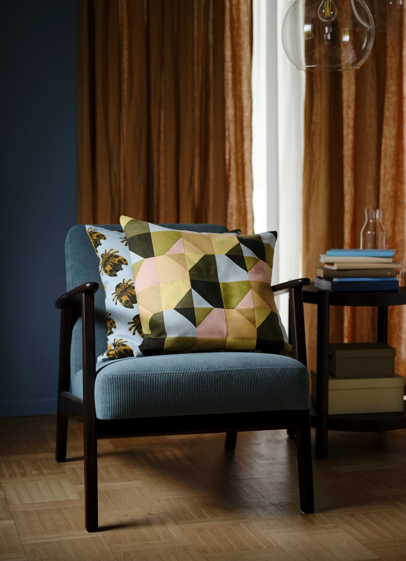A grey-turquoise armchair in front of dark beige curtains with a cushion inside a SVARTHÖ multicoloured cushion cover on it.