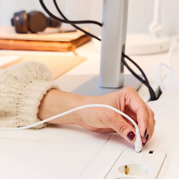 Eine Person steckt ein USB-Kabel in eine weiße KOPPLA 4-fach-Steckdose mit 2 USB-Anschlüssen, die auf einem MITTZON Sitz-/Stehschreibtisch liegt.