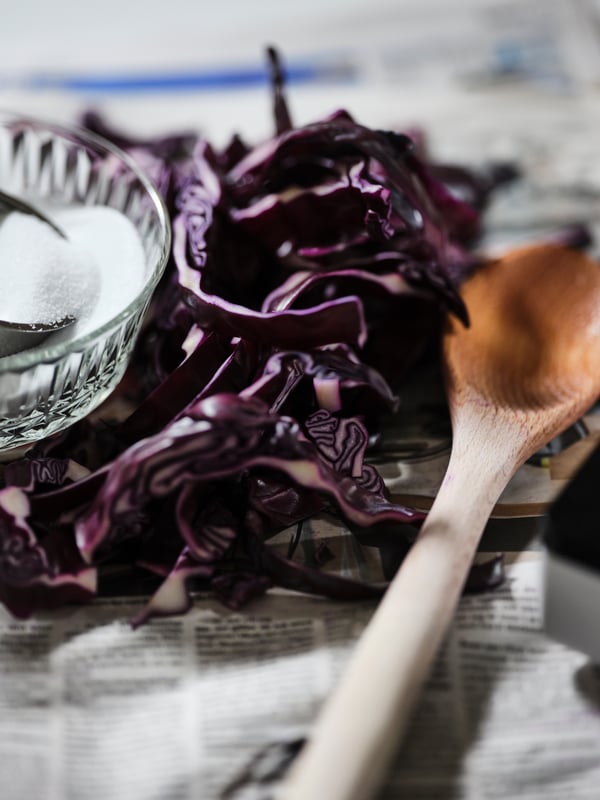 A round RÖRT beech spoon is placed alongside sliced red cabbage and a glass bowl of salt on a spread newspaper.