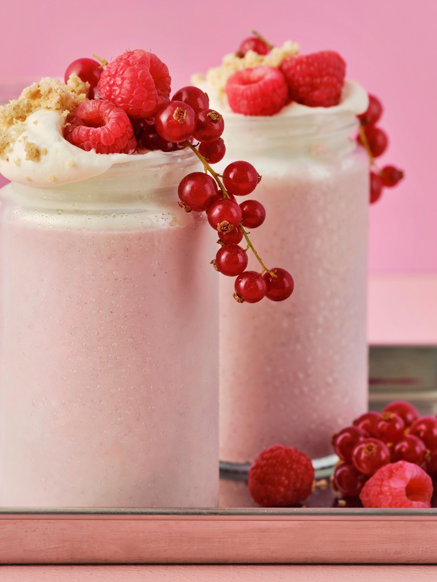 Two light pink LÅNGLUR fruit smoothie milkshakes are topped with red raspberries, red berries, and cookie crumbs.