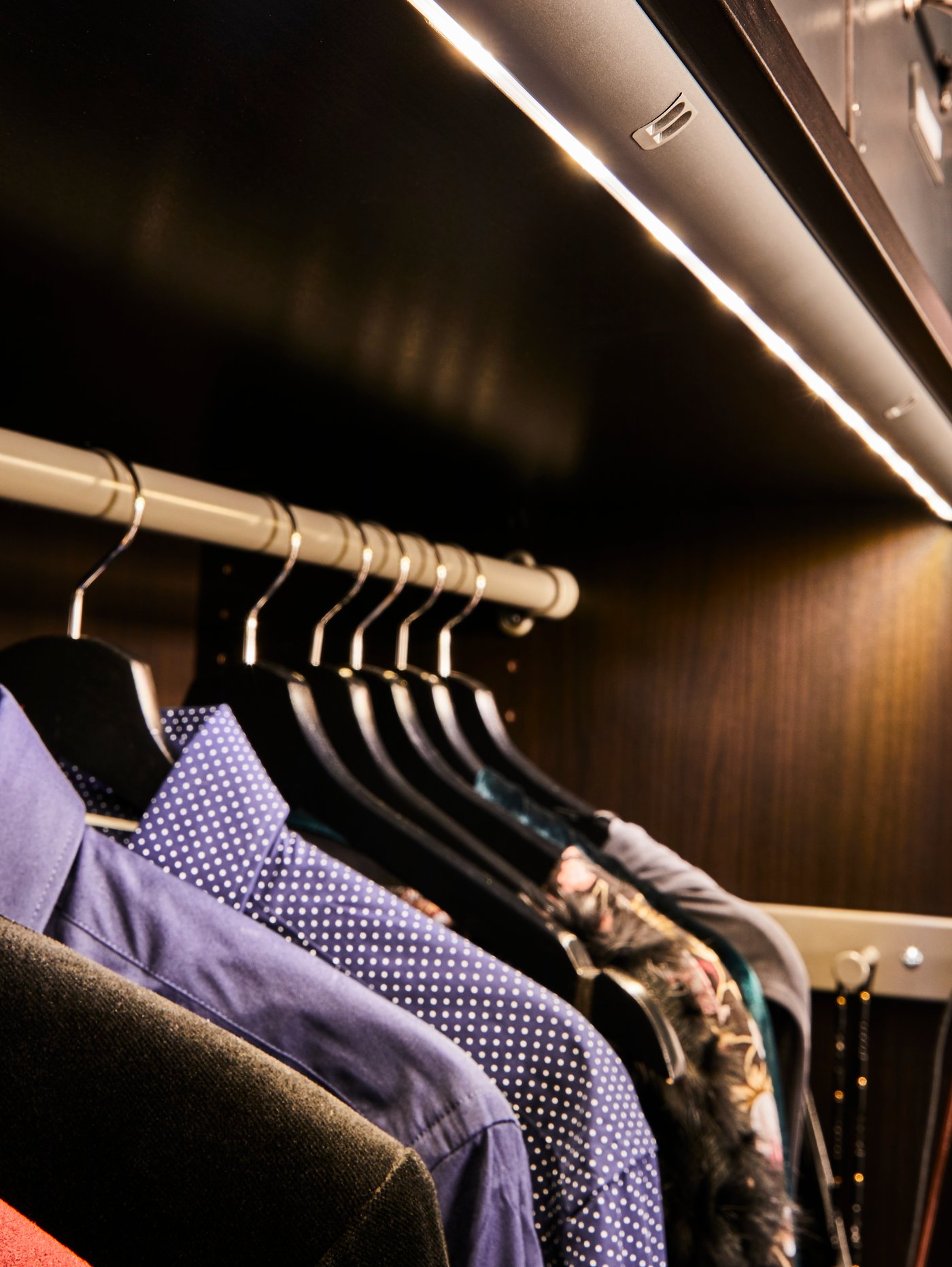 Integrated lighting as part of a storage unit, with various coloured shirts hanging from a clothes rail by coat hangers.