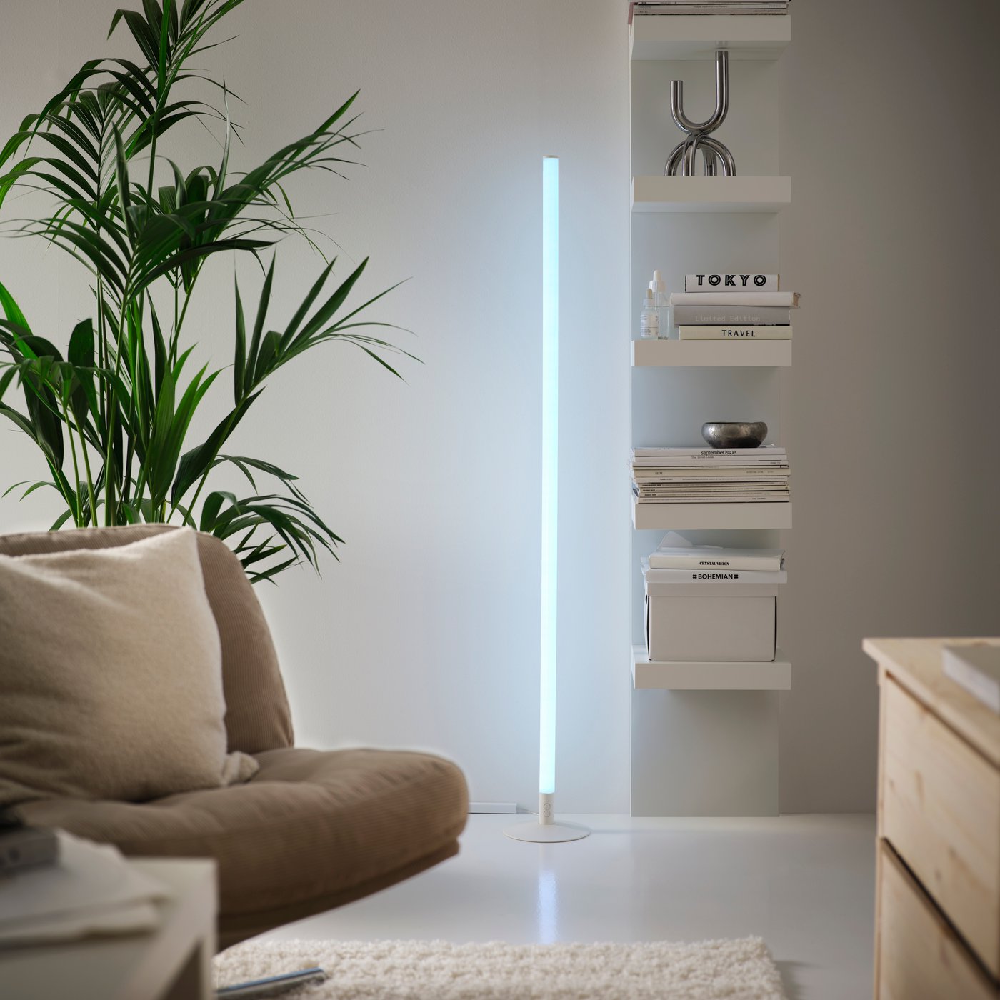 FÖRLIG LED floor/wall lamp with light blue glow, standing next to a wall shelf unit with a SNÖDUVA candlestick.