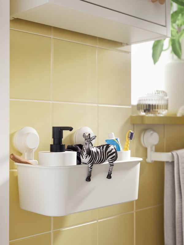 Dozator za sapun, četkica za zube, pasta za zube i igračka zebra u beloj TISKEN korpi s vakuumskim držačem na zidu sa žutim pločicama.