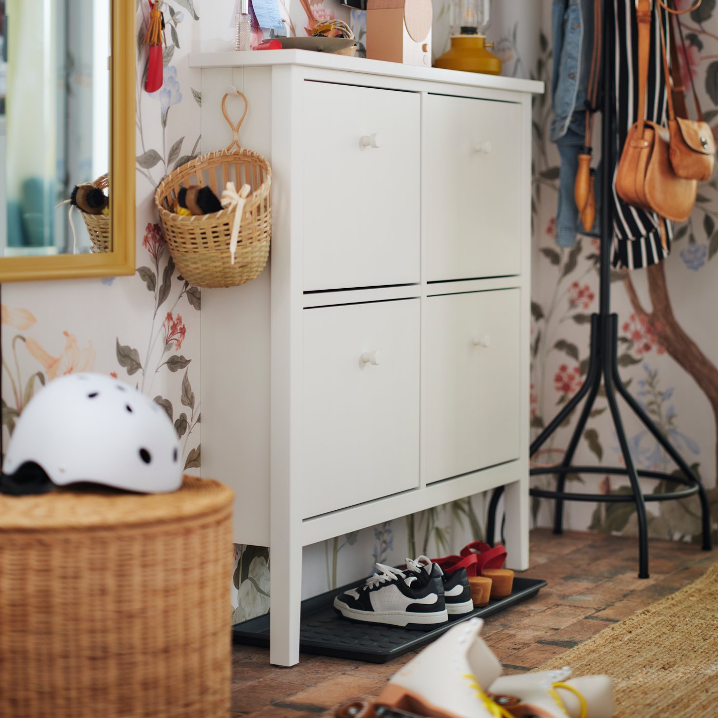 A white GULLABERG shoe cabinet against a floral wall with a basket hanging on one side and a BAGGMUCK shoe tray underneath.