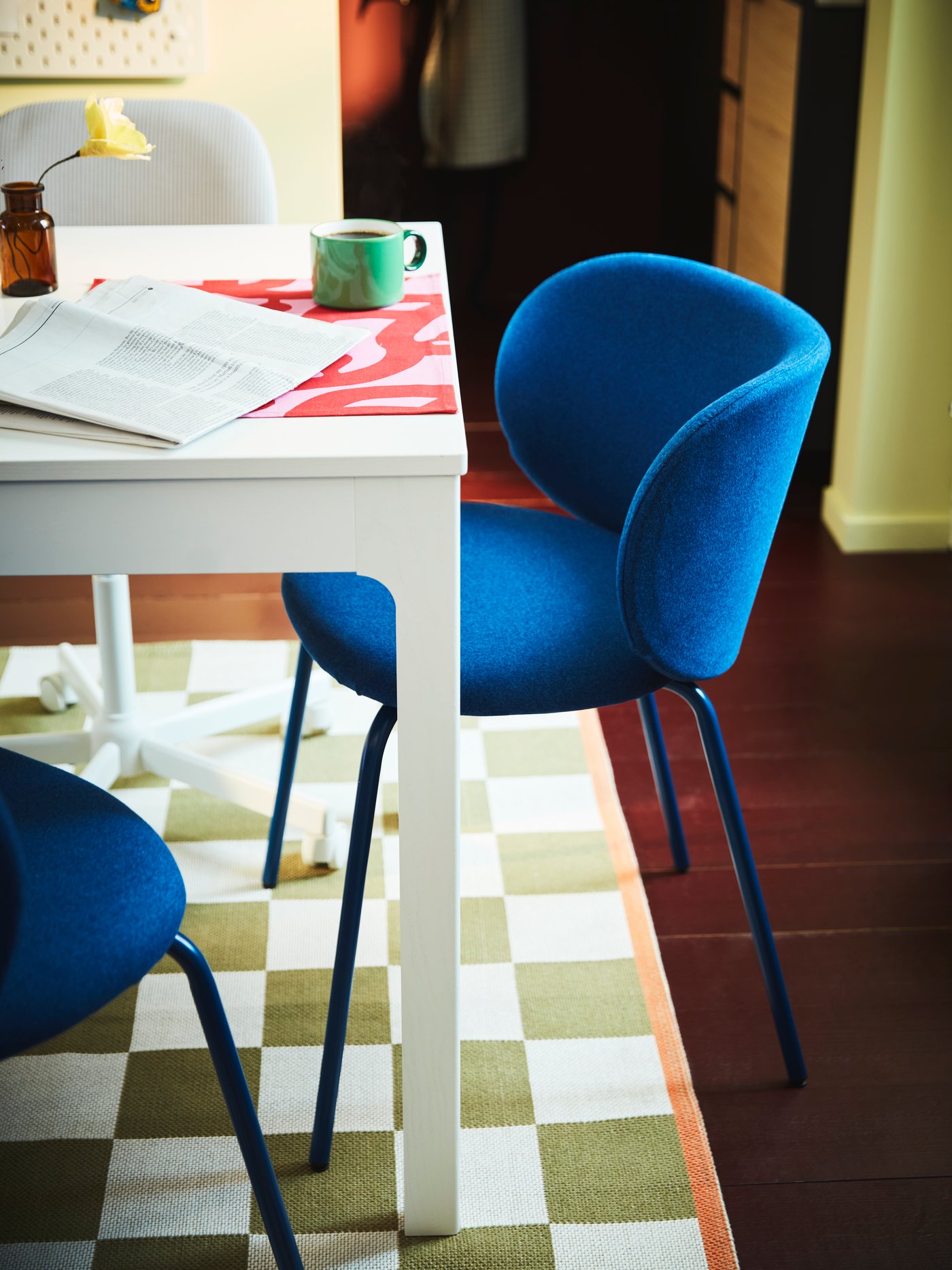 Two blue KRYLBO chairs stand on a white and green patterned rug, by a white table with a green mug and an open newspaper.