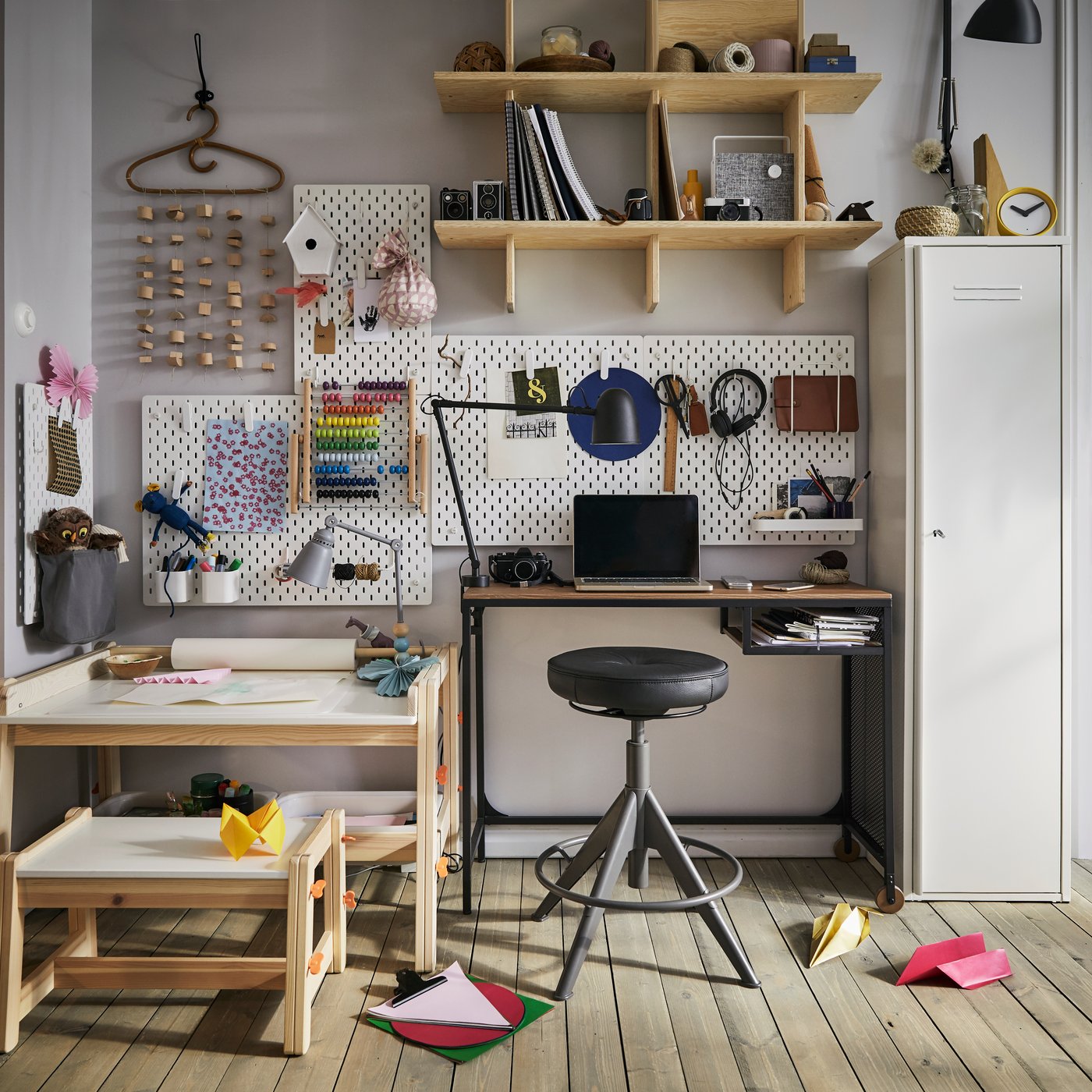 Ein Kindertisch mit Bastelutensilien und ein Erwachsenenschreibtisch mit aktiver TROLLBERGET Sitz-/Stehstütze mit Bezug „Glose“ in Schwarz und einer Lochplatte an der Wand.