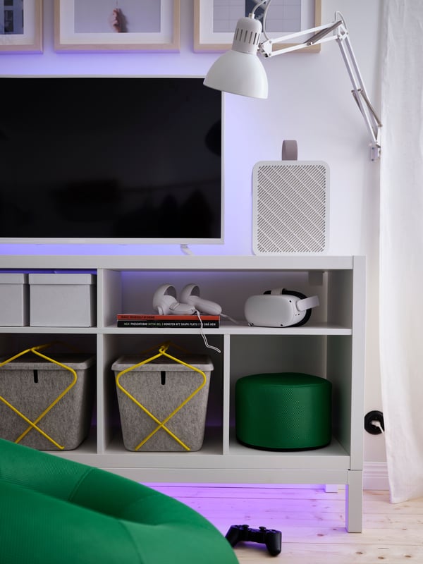 Una tira de iluminación LED ORMANÄS oculta emite un resplandor púrpura debajo y detrás de una consola de juegos sobre un banco de TV blanco.