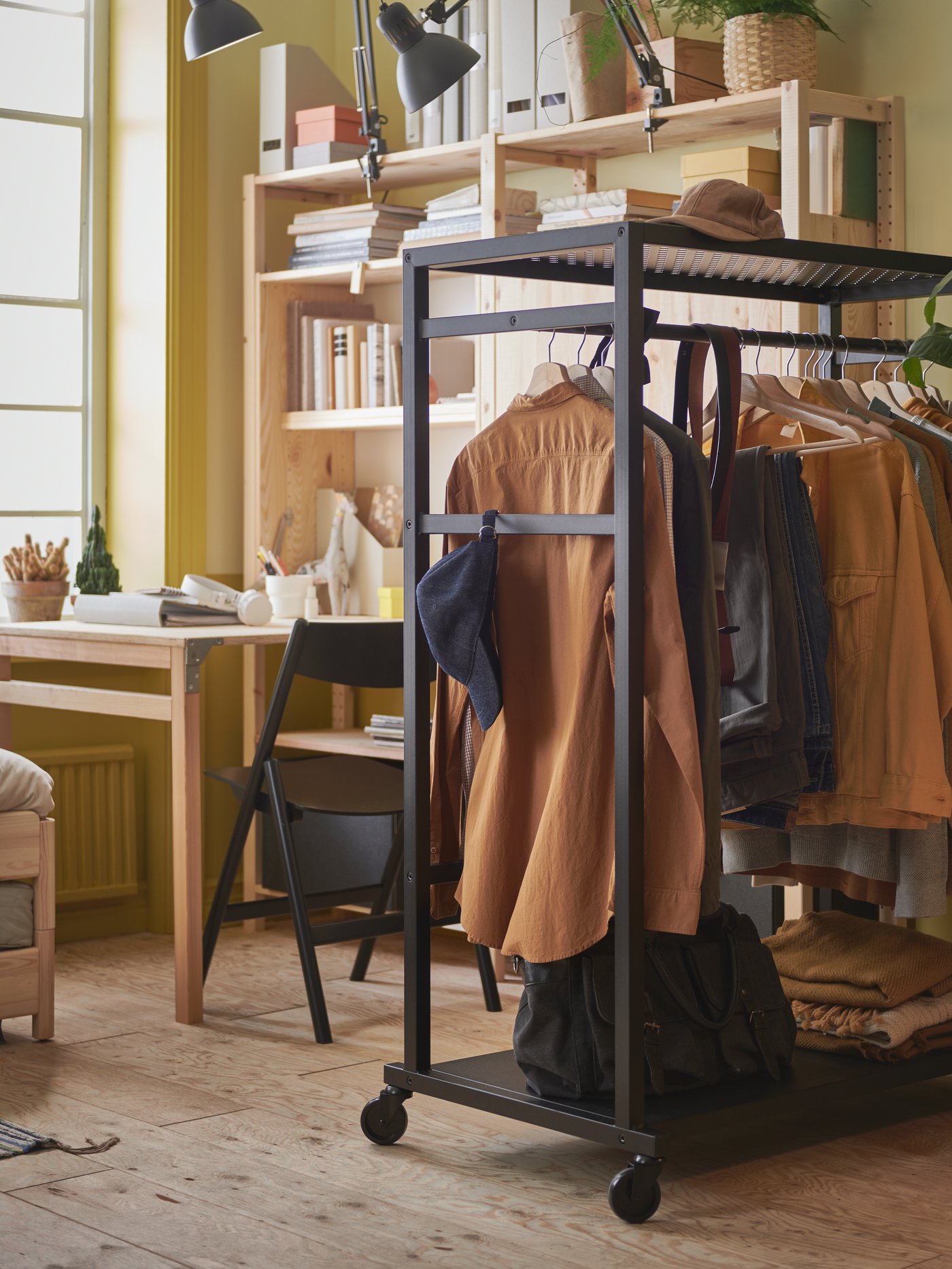 A student dorm room with a black open storage unit on wheels with hanging clothes, and a desk and bookcase in the background.